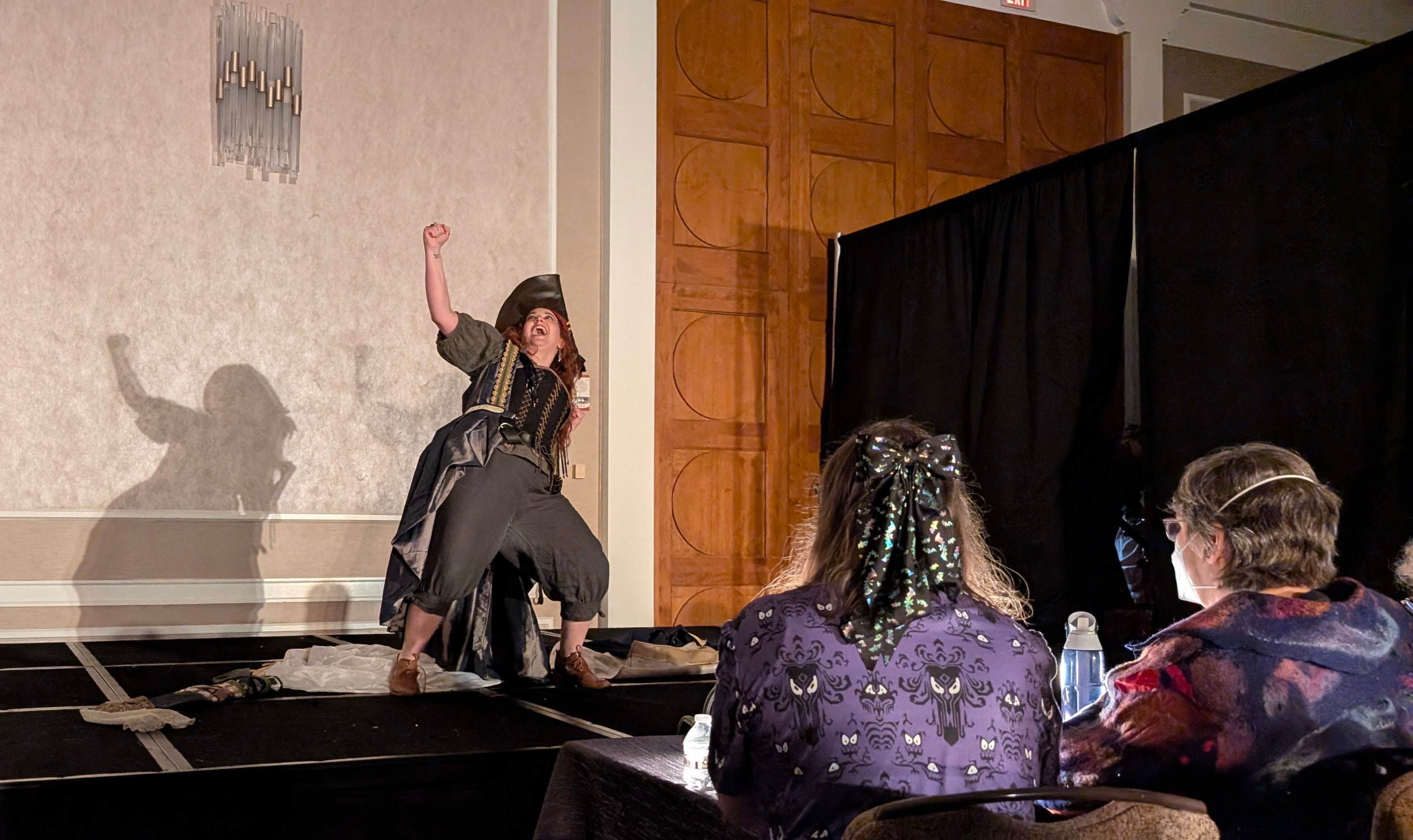 This is "The Princess Problem," which won best in show. The stage of the Arisia Masquerade. A woman in a corset and pirate hat raises her fist in exultation. Her mouth is open in a scream of joy. On the lower right, two judges observe the costume's wearer.